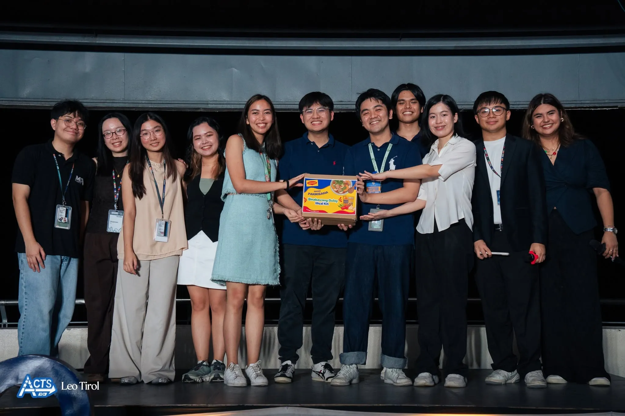 The ACTS 2025 team and event organizers posing onstage while presenting a Maggi package.