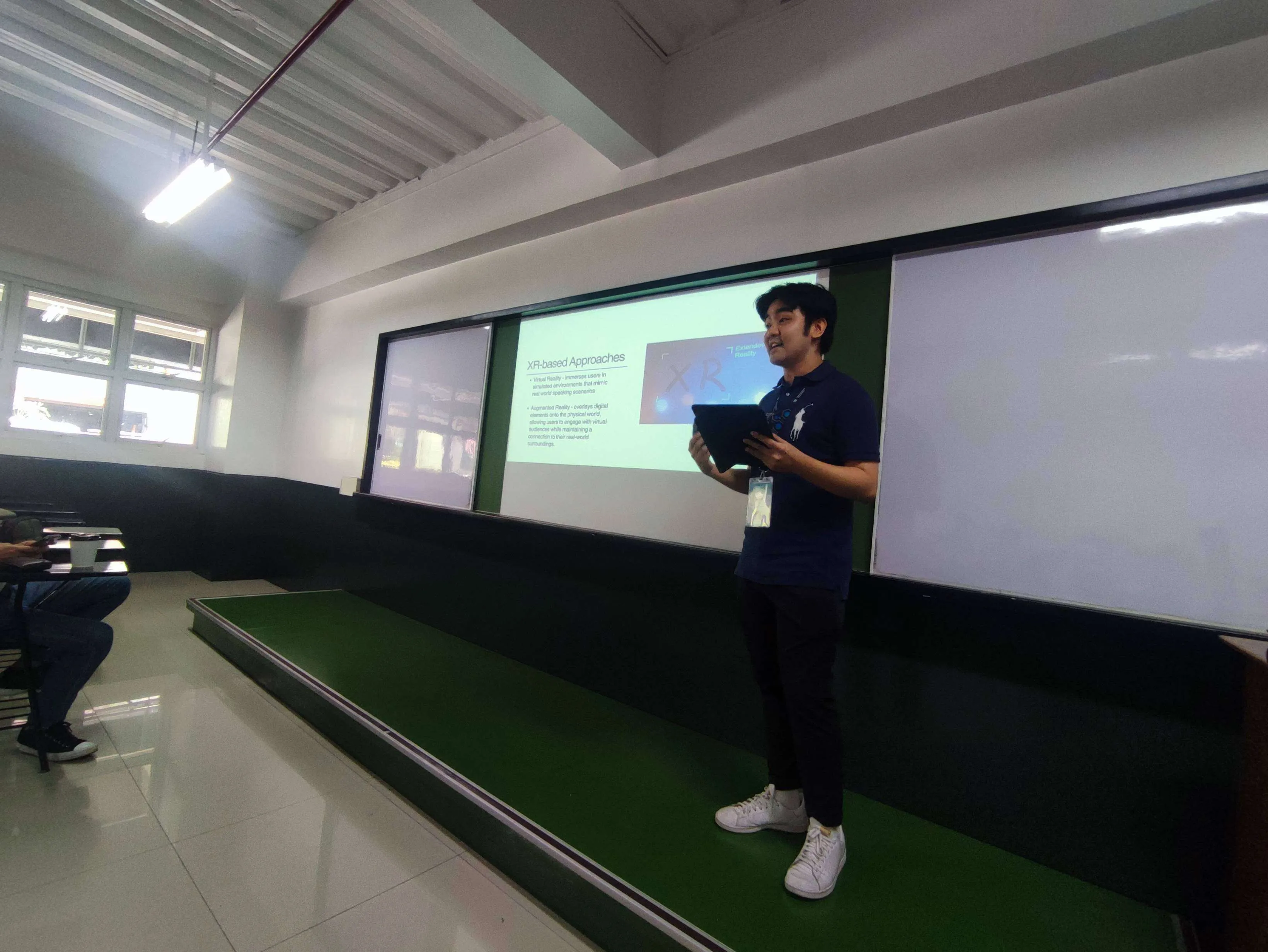 Jan Benjamin Yap presenting SpeakAR on a lecture stage, holding a tablet running the app while a slide titled "XR-based Approaches" plays behind him.